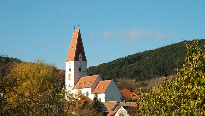 Pfarrkirche Nußdorf, © Blesl Pfarrkirche Nußdorf mit rotem Dach und Uhrturm, umgeben von Bäumen und Hügeln.