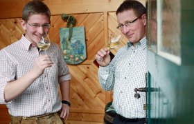 Fam. Baumgartner, © weinfranz.at Zwei Männer in karierten Hemden trinken Weißwein in einem holzgetäfelten Raum.