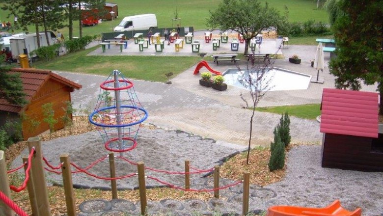 Waldbad-Spielplatz, © Marktgemeinde Wölbling Spielplatz mit Klettergerüst, Rutsche und kleinem Pool, umgeben von Bäumen und Wiese.