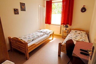 Zimmeransicht, © Martin Herget Ein einfach eingerichtetes Zimmer mit zwei Einzelbetten, einem Fenster mit roten Vorhängen und einem kleinen Tisch mit Stuhl.