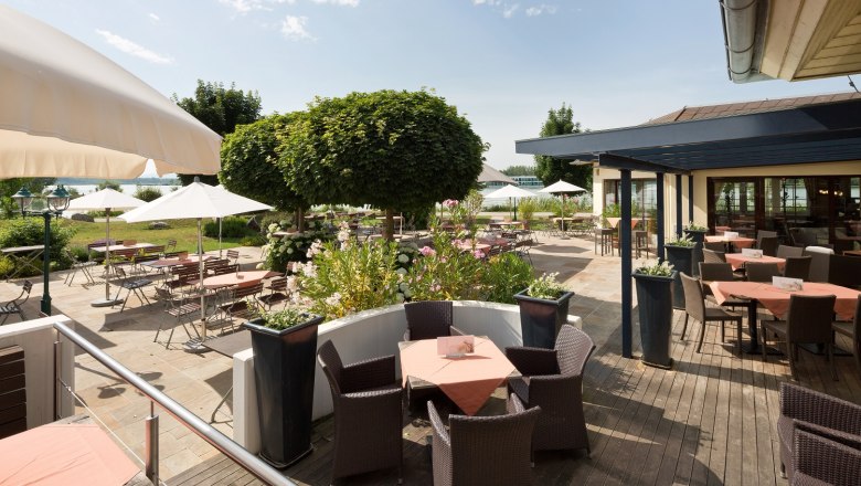 Terrasse, © DR Sonnige Terrasse mit Tischen, Stühlen und Sonnenschirmen, umgeben von Pflanzen und Bäumen.