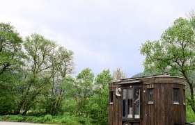 TD-Wohnwagon Karl aussen 2, © WohnwagonGmbH Ein kleiner, dunkler Wohnwagen mit Holzverkleidung steht auf einem Parkplatz vor grünen Bäumen und bewölktem Himmel.