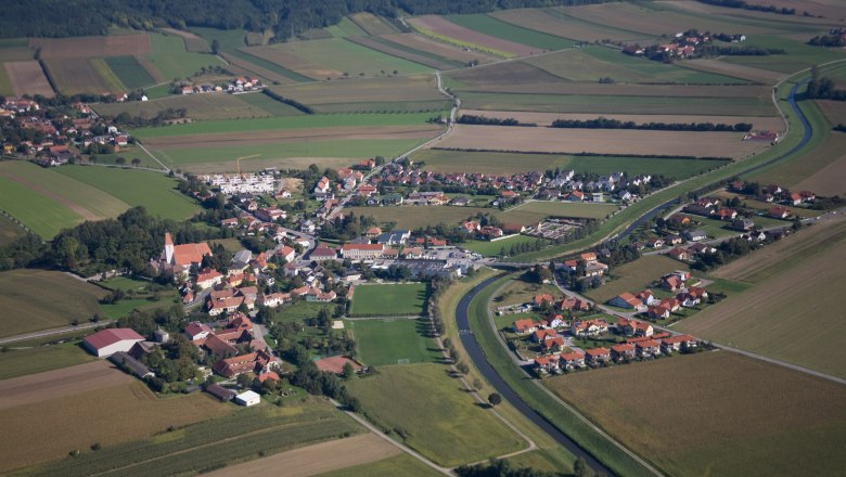 von oben, © Irene Köszali Luftaufnahme eines Dorfes mit umliegenden Feldern und einem Fluss.