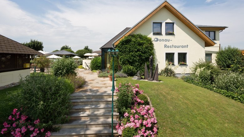 Haus, © DR Ein Restaurant mit Garten und Terrasse, umgeben von Blumen und Büschen.