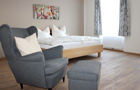 Schlafzimmer, Bett & Rad Schöpf, Traismauer, © Familie Schöpf Grauer, gemütlicher Stuhl im Vordergrund. Im Hintergrund ein Doppelbett mit weißer Bettwäsche.