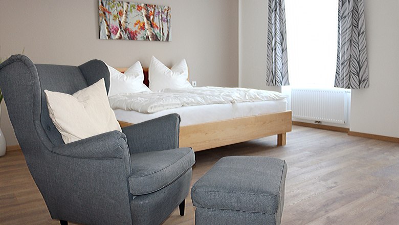 Schlafzimmer, Bett & Rad Schöpf, Traismauer, © Familie Schöpf Grauer, gemütlicher Stuhl im Vordergrund. Im Hintergrund ein Doppelbett mit weißer Bettwäsche.