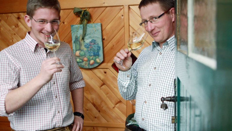 Zwei Männer in karierten Hemden trinken Weißwein in einem holzgetäfelten Raum.