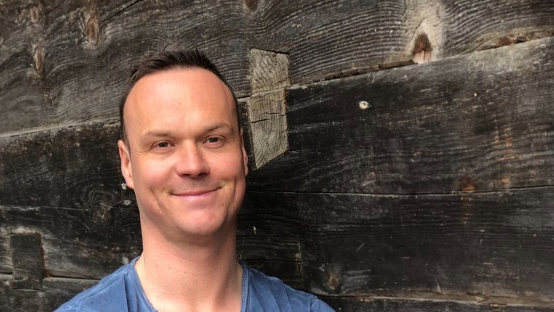 Mann in blauem T-Shirt vor Holzwand.