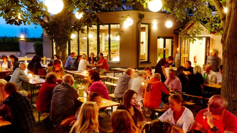 Menschen sitzen abends an Tischen im Freien, umgeben von Bäumen und Lichtern.