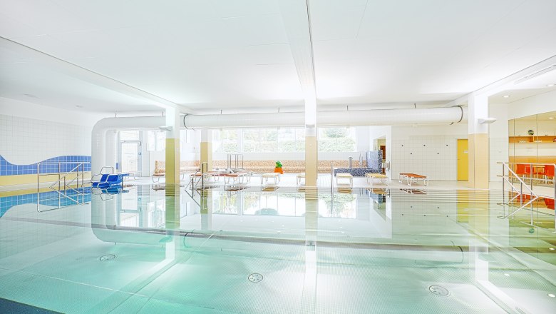 Innenansicht eines hellen Hallenbads mit Schwimmbecken, Liegestühlen und großen Fenstern.