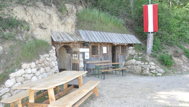 Eine rustikale Holzhütte mit Tisch und Bänken davor, eingebaut in einen Hügel. Eine rot-weiße Fahne hängt an einem Baum.