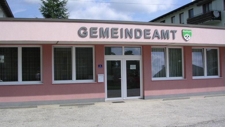 Außenansicht eines rosa Gebäudes mit der Aufschrift 'GEMEINDEAMT' und einem Wappen.