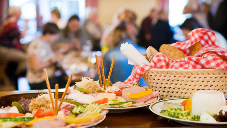 Ein Tisch mit traditionellen österreichischen Speisen, darunter Aufschnitt, Käse und Brot in einem Korb mit rot-weiß kariertem Tuch.