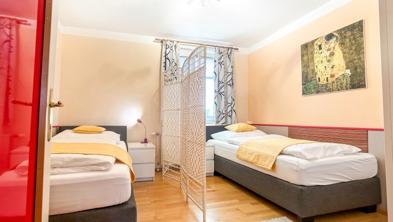Schlafzimmer mit zwei Einzelbetten, gelber Bettwäsche, einem Raumteiler und einem Gemälde an der Wand.