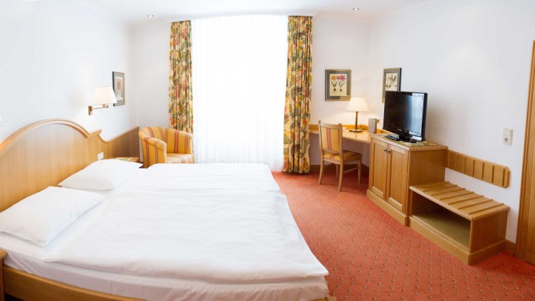 Ein Hotelzimmer mit Doppelbett, Schreibtisch, Fernseher und Sessel. Helle Vorhänge und florale Vorhänge schmücken das Fenster.