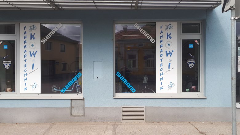 Fahrradgeschäft mit blauer Fassade und Schaufenstern mit Aufschrift 'Fahrradtechnik KOWI'.