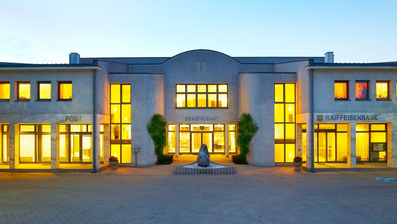 Beleuchtetes Gebäude mit Aufschrift 'Gemeindeamt', 'Post' und 'Raiffeisenbank'.