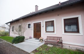 Eingang einer rosa gestrichenen Ferienwohnung mit Holzt&uuml;r, zwei Fenstern und einer Holzbank daneben.