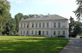Schloss Würmla, ein zweistöckiges Gebäude mit grauem Dach und weißen Wänden, umgeben von Bäumen und Rasen.