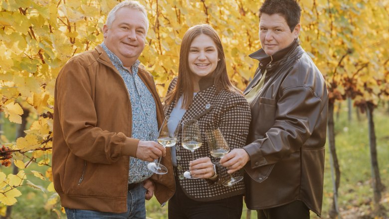 Drei Personen stoßen mit Weingläsern in einem herbstlichen Weinberg an.