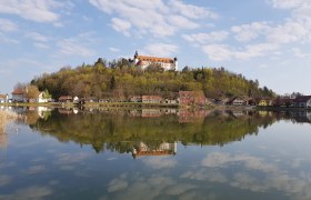 Ein Schloss auf einem Hügel spiegelt sich in einem ruhigen See, umgeben von Bäumen und kleinen Häusern.