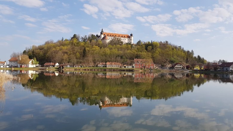 Ein Schloss auf einem Hügel spiegelt sich in einem ruhigen See, umgeben von Bäumen und kleinen Häusern.