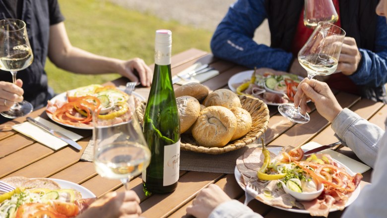 Menschen genießen Wein und Jause im Freien an einem Holztisch.
