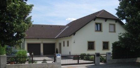 Ein modernes Einfamilienhaus mit braunem Dach und hellem Anstrich, umgeben von B&auml;umen und einem Zaun.