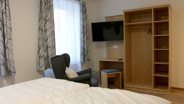 Offener Kleiderschrank mit Fernseher an der Wand. Vor einem Fenster mit einer Gardine steht ein gr&uuml;ner gem&uuml;tlicher Stuhl.