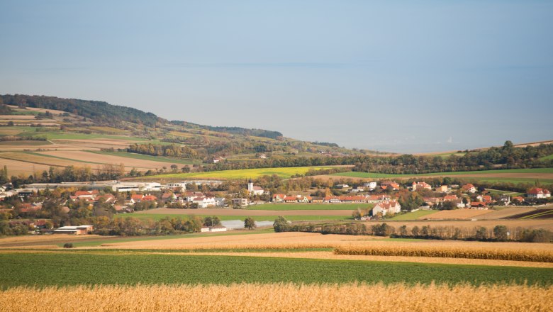 Landschaft mit Feldern und einem Dorf im Hintergrund, umgeben von H&uuml;geln und B&auml;umen.