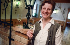 Eine lächelnde Person hält ein Glas Weißwein in einem gemütlichen Raum mit Backsteinmauer und Laterne.