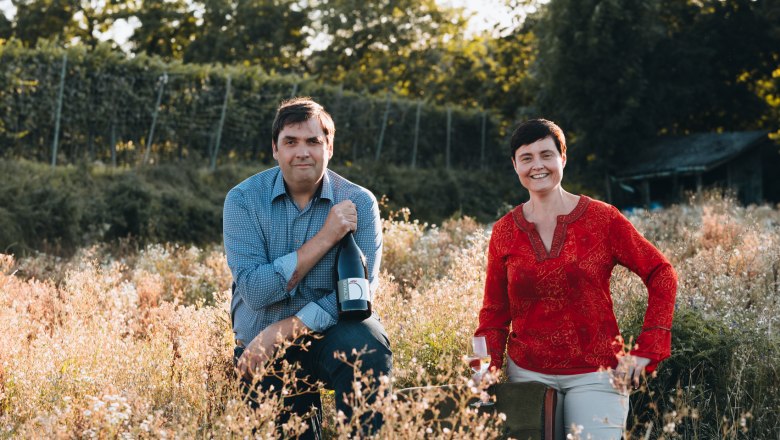 Zwei Personen in einem Weinberg, eine hält eine Weinflasche, die andere ein Weinglas.