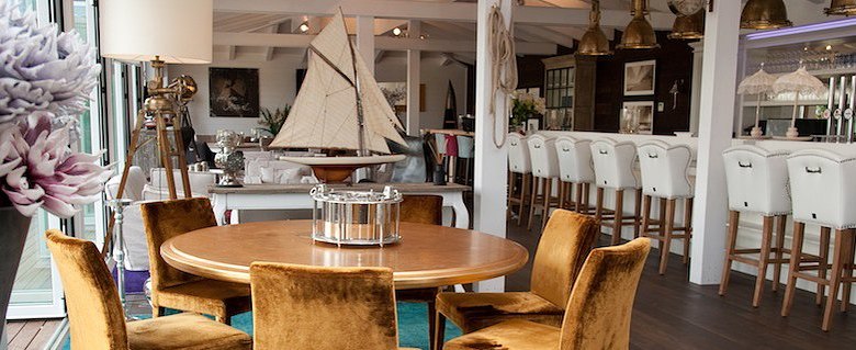 Eleganter Barbereich mit runden Holztischen, samtigen Stühlen und einer Bar mit weißen Hockern.