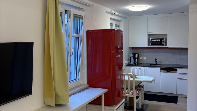 Modernes Wohnzimmer mit offener Küche, rotem Kühlschrank, gelben Vorhängen und Holzboden.