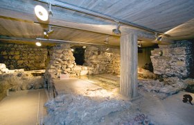 Unterirdische Ruinen mit Steinmauern und Betonstützen, beleuchtet durch Deckenstrahler.