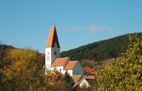 Kirche in Nußdorf mit rotem Dach und Uhrturm, umgeben von Bäumen und Hügeln.