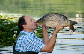 Ein Mann küsst einen Karpfen am Ufer eines Sees mit Seerosen.