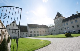 Eingang von Schloss Walpersdorf mit offenem Tor, Rasenfläche und historischem Gebäude im Hintergrund.