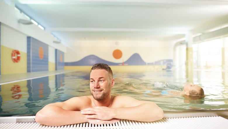 Ein Mann lehnt sich am Beckenrand eines Hallenbads an, während im Hintergrund eine Person im Wasser schwimmt.