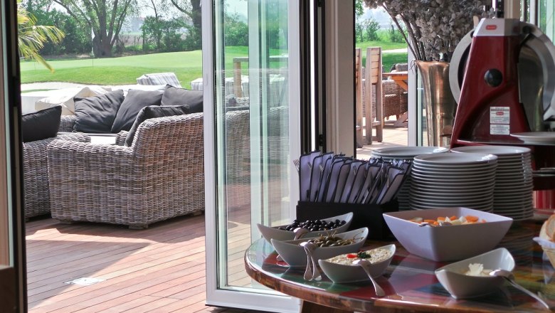 Ein gedeckter Tisch mit Geschirr und Snacks, daneben eine Terrasse mit Rattanmöbeln und Blick auf einen grünen Rasen.