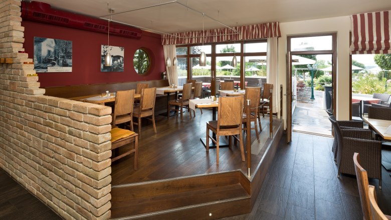 Ein gem&uuml;tliches Restaurant mit Holzm&ouml;beln, roten W&auml;nden und gro&szlig;en Fenstern, die auf eine Terrasse f&uuml;hren.