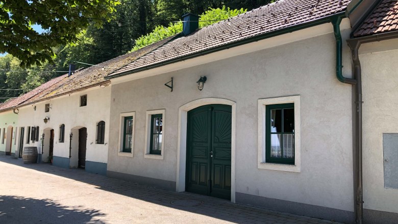 Außenansicht eines traditionellen Heurigen mit weißen Wänden und grünem Tor, umgeben von Bäumen.