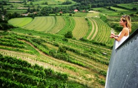 Frau mit Weinglas blickt von einem Aussichtspunkt auf grüne Weinberge.