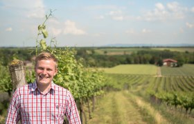 Ein lächelnder Mann steht in einem Weinberg mit weitem Blick über die Landschaft.