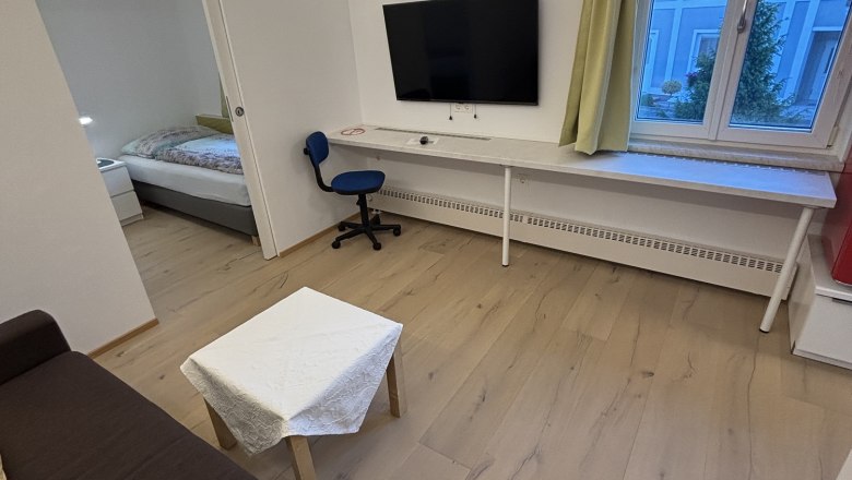 Modernes Wohnzimmer mit braunem Sofa, kleinem Tisch, Fernseher an der Wand, Schreibtisch und Stuhl. Fenster mit Blick auf ein blaues Gebäude.