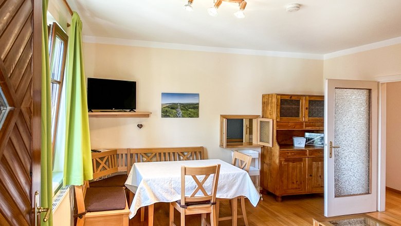 Esszimmer mit Holzm&ouml;beln, Tisch mit wei&szlig;er Tischdecke, gr&uuml;nen Vorh&auml;ngen und Fernseher an der Wand.