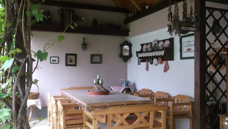 &Uuml;berdachte Terasse mit Tisch und St&uuml;hlen, im Hintergrund eine Wand mit verschiedener Wanddeko.