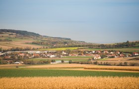 Landschaft mit Feldern und einem Dorf im Hintergrund, umgeben von Hügeln und Bäumen.