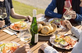 Menschen genießen Wein und Jause im Freien an einem Holztisch.